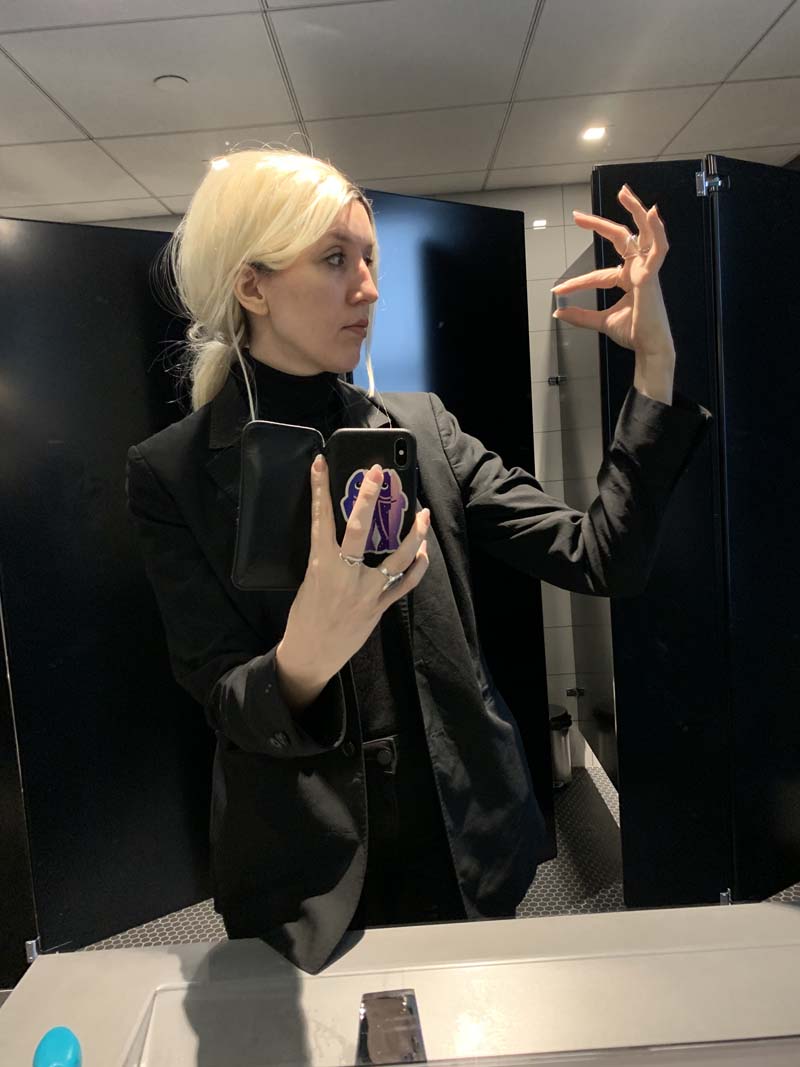 me with a blonde wig in a bathroom, wearing all black and holding up a pill-like object and looking at it