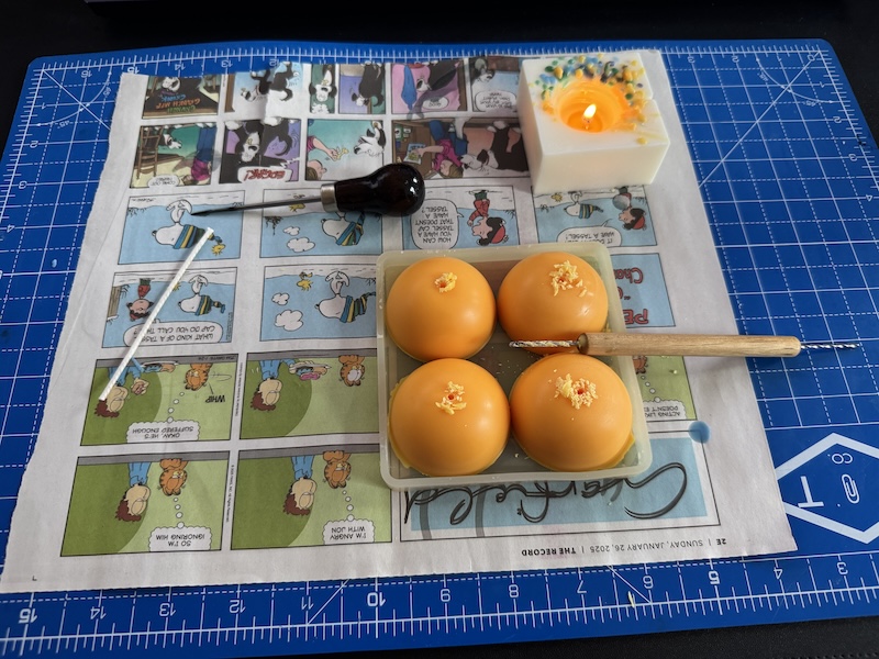photo of a cutting mat on my desk and on it i am wicking "egg yolk" like candles. i have a little hand drill and an awl, as well as a small candle that i'm using to heat the tools. everything is on a page from the sunday comics to protect the mat
