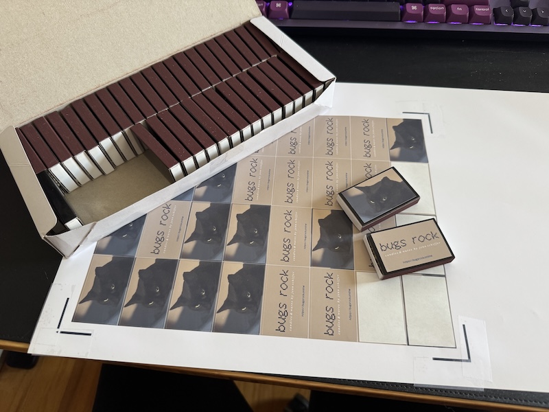 photo of my desk with a sheet of sticker labels of pumagreg and the bugs rock brand that are on a couple matchboxes, next to them is a box of "blank" matchboxes