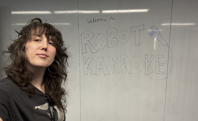 selfie of me in front of a classroom white board that says "welcome to robot karaoke"