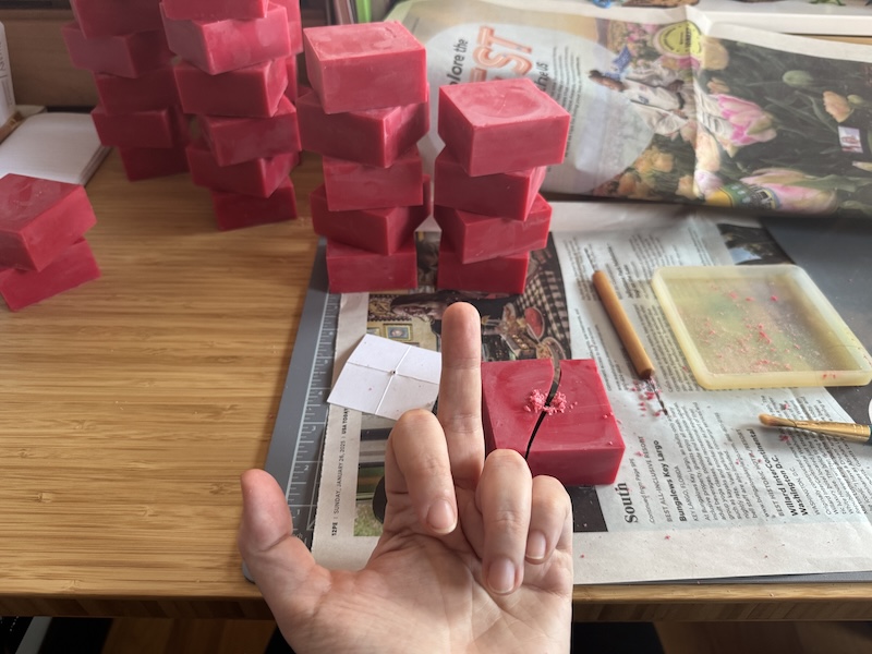 photo of my hand giving the middle finger to a red square candle i broke while drilling a whole in it for a wick, behind it are over a dozen of the same kind of candle that i successfully drilled holes in without breaking