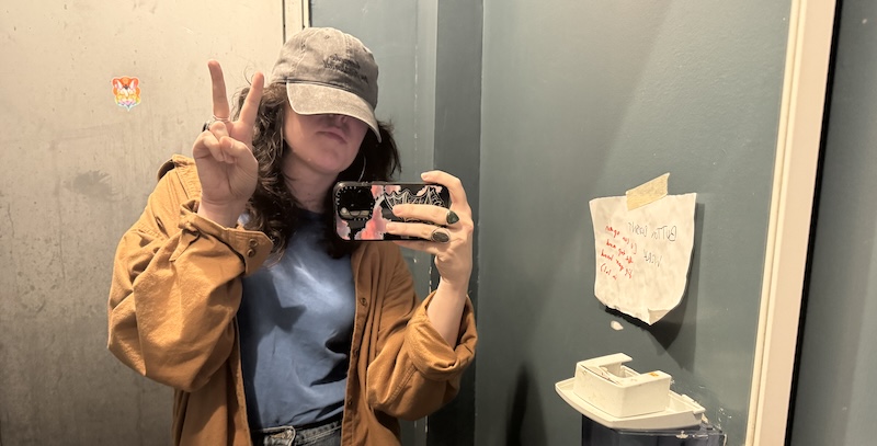 bathroom selfie of me at the venue where we did karaoke, i've covered most of my face with my "karaoke professional" baseball cap. there's a sign on the wall.