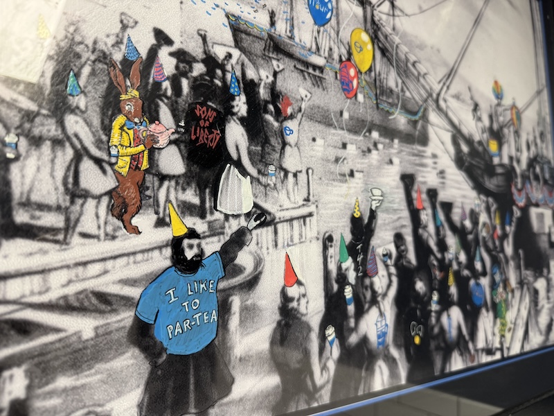 photo of an drawing (on the wall of a coffee shop in boston) of the boston tea party and paint marker is used to give the people party hats, and one person is wearing a shirt that says "i like to par-tea"