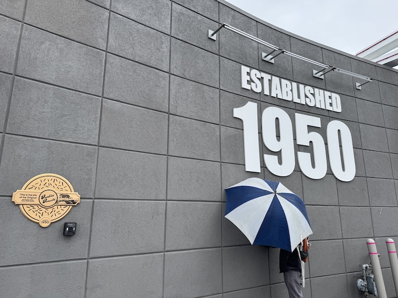 photo of the first ever dunkin that says "established 1950" and mike is using himself and the umbrella to hide an obnoxious instagram logo. there's a plaque that says this is the site of the original dunkin