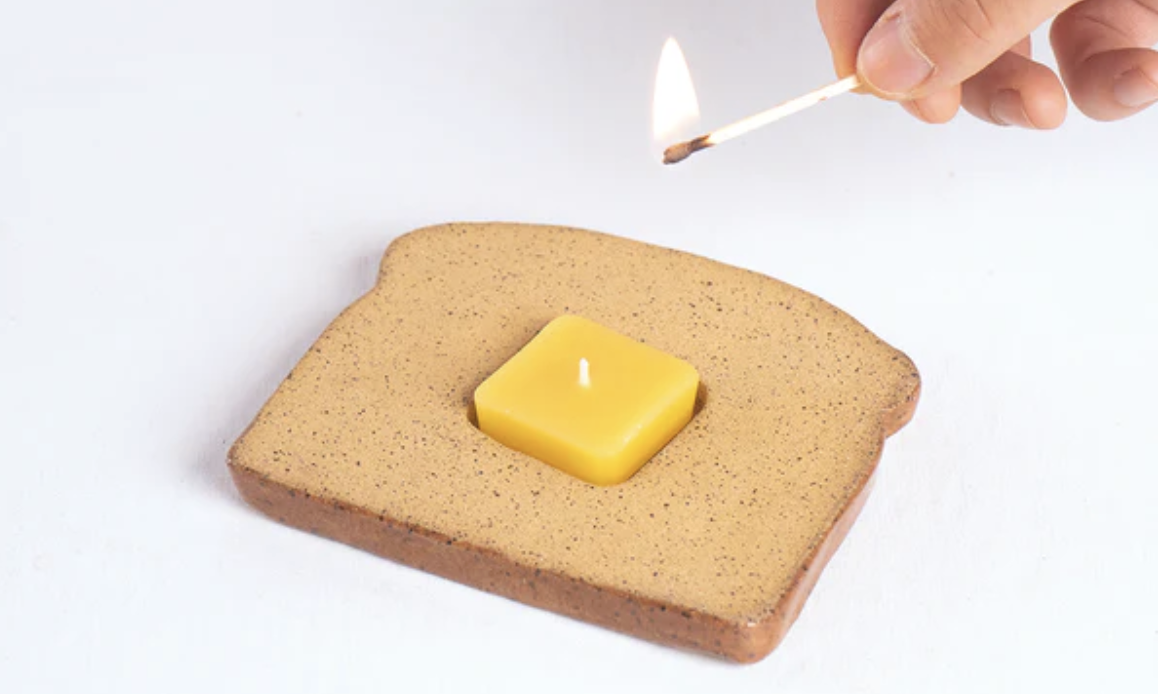 product photo of a ceramic piece of toast with a tab of butter candle made of beeswax about to be lit by a hand holding a lit match