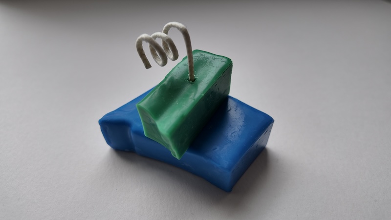 photo of a candle i made on a white backdrop. the candle has a base of half a broken square of blue and on top of it is half a broken thin rectangle of green