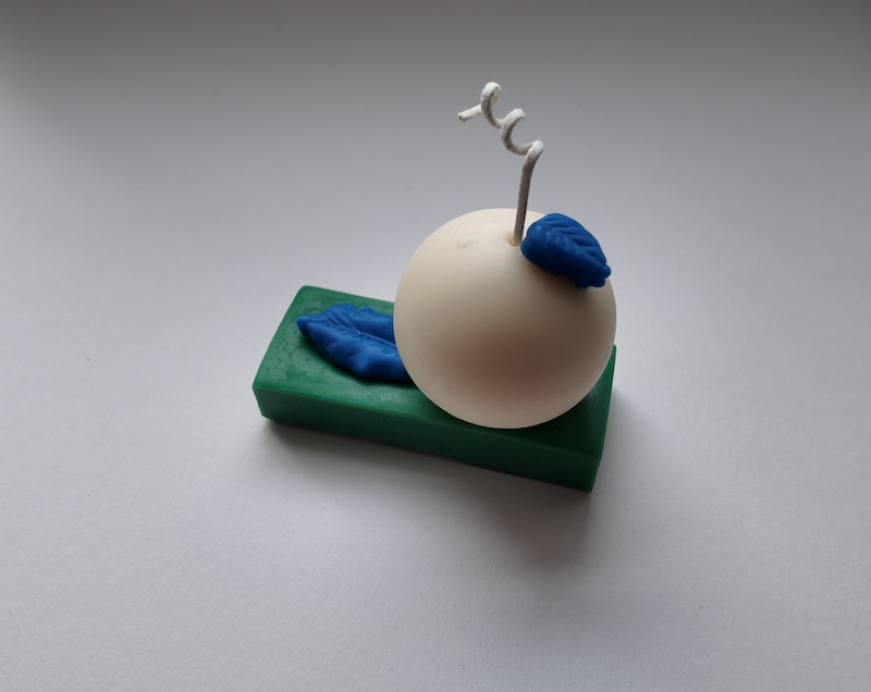 photo of a candle i made on a white backdrop. the candle has a green rectangular base with a white sphere on it and two little blue leaves