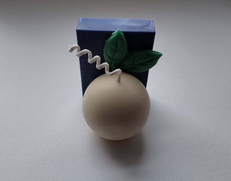 photo of a candle i made on a white backdrop. the candle has a base of a white sphere with a purple rectangle alongside the back of it and a couple of green leaves
