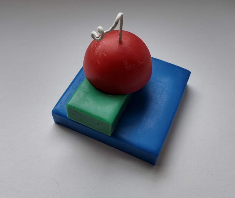 photo of a candle i made on a white backdrop. the candle has a blue square base, a green rectangle on top of it, and then a red half-sphere on top of the green piece
