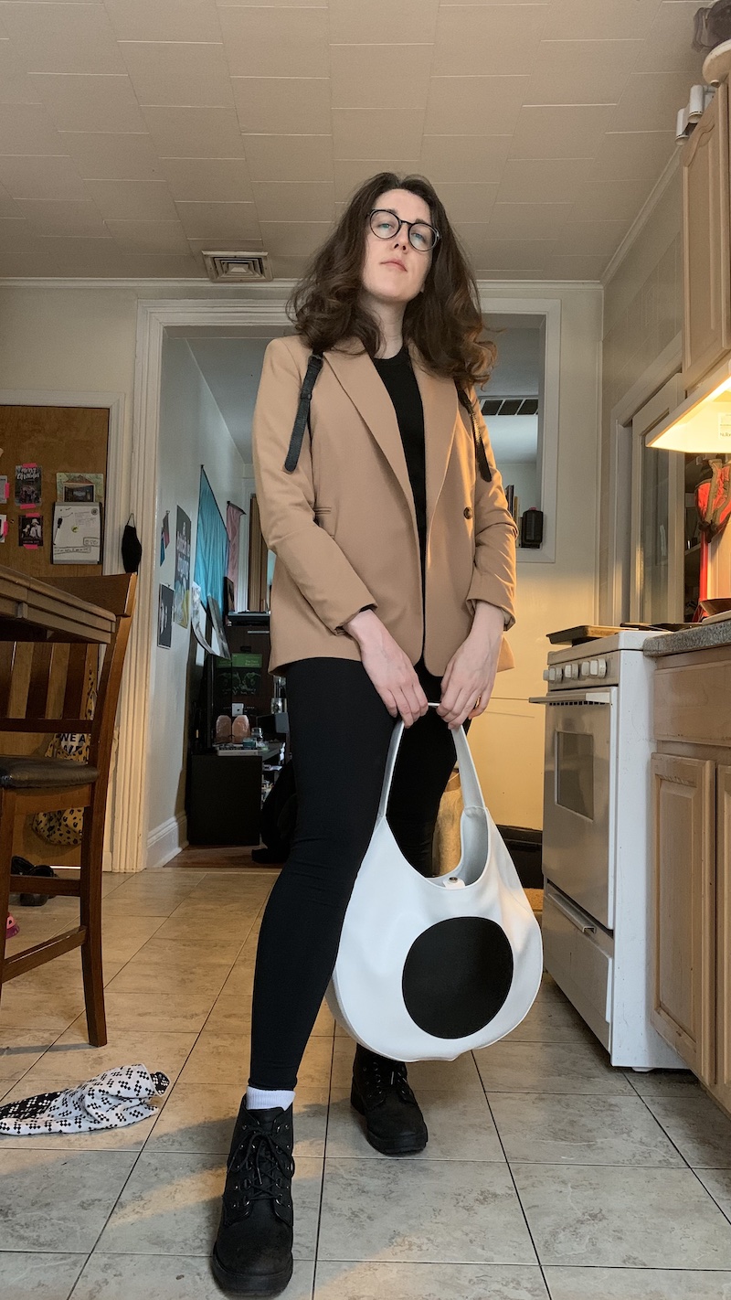 photo of me standing in my kitchen and posing with a white and black bag, wearing a tan blazer and black leggings and boots