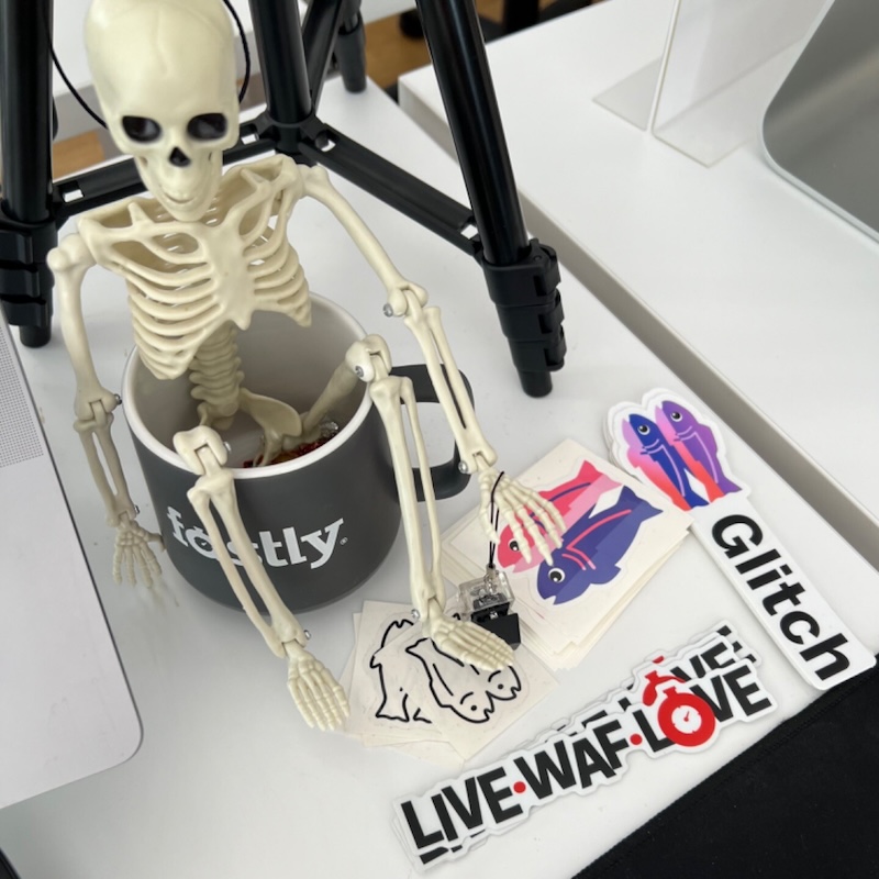 photo of my work desk with a skeleton figure sitting in a fastly mug and around it are glitch and fastly stickers