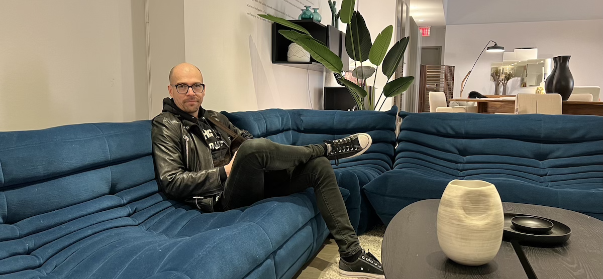 photo of juan pablo, a handsome bald colombian man in leather, sitting on a large expensive blue sectional in a high-end furniture store