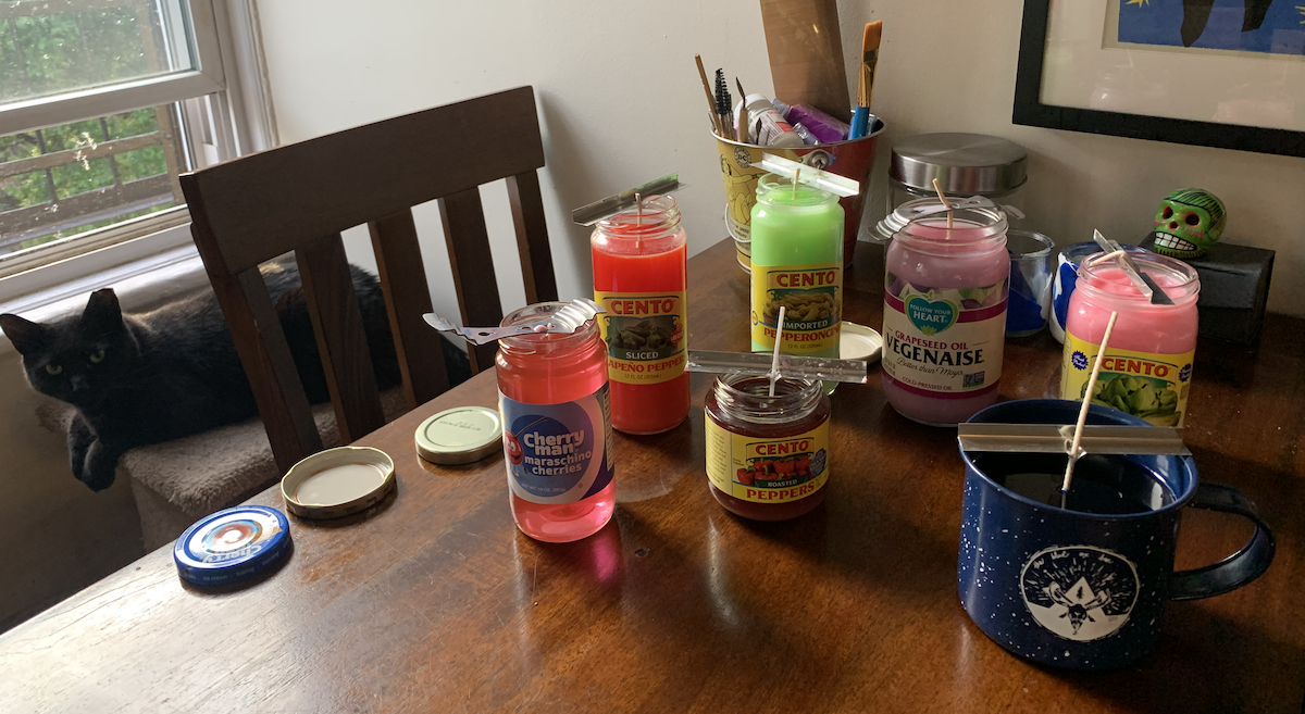 a photo of my kitchen table, with my black cat pumagreg in the background, covered in jar lids and newly-poured colorful candles