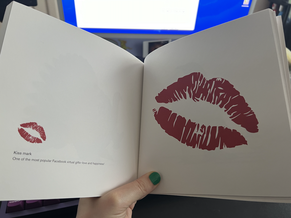 photo of me holding up susan kare's "icons" book opened up to the page of the facebook kiss mark "one of the most popular facebook virtual gifts--love and happiness" that's similar but very different in texture and color and not rotated as much as the maxos icon