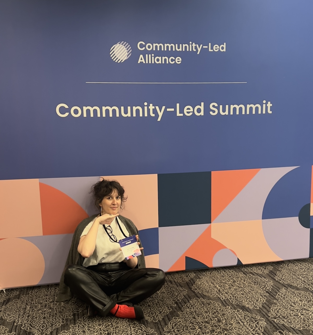photo of me on the floor in front of a large banner that says community-led alliance, community-led summit in a drab conference room but i'm posing all cute, hand under chin like i'm a sweet angel