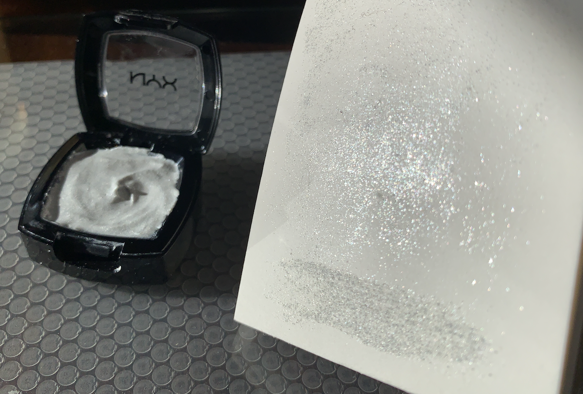 photo of my new paint in its pan and i'm holding up a small paper swatch of the paint, it's more a silver micro glitter than a pigmented color