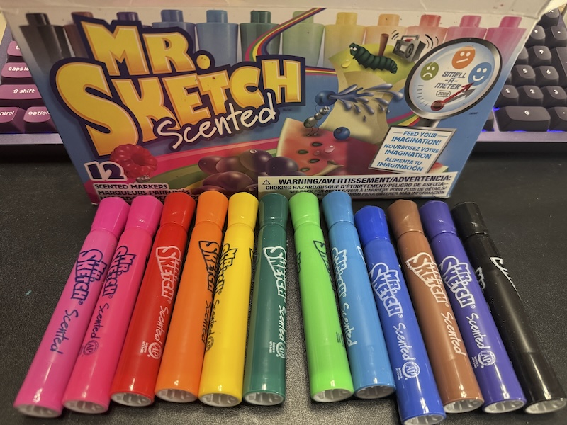 photo of my desk with a 12 pack of dr. sketch scented markers and i have the markers laid out in front of the box. the colors are standard colors from the rainbow with additional pink, brown and black