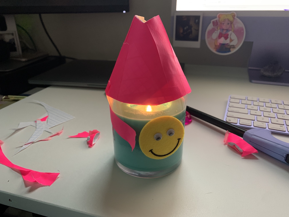 my candle with a cone hat made of paper and hot pink tape, the desk around it covered in paper shrapnel from my cutting it all up