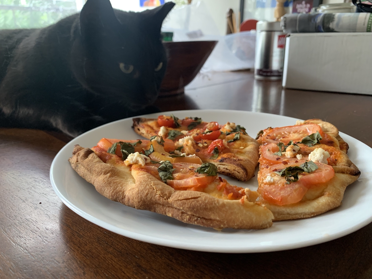 photograph of a plate with two slices of tomato flatbread and a black cat on the table looking at it