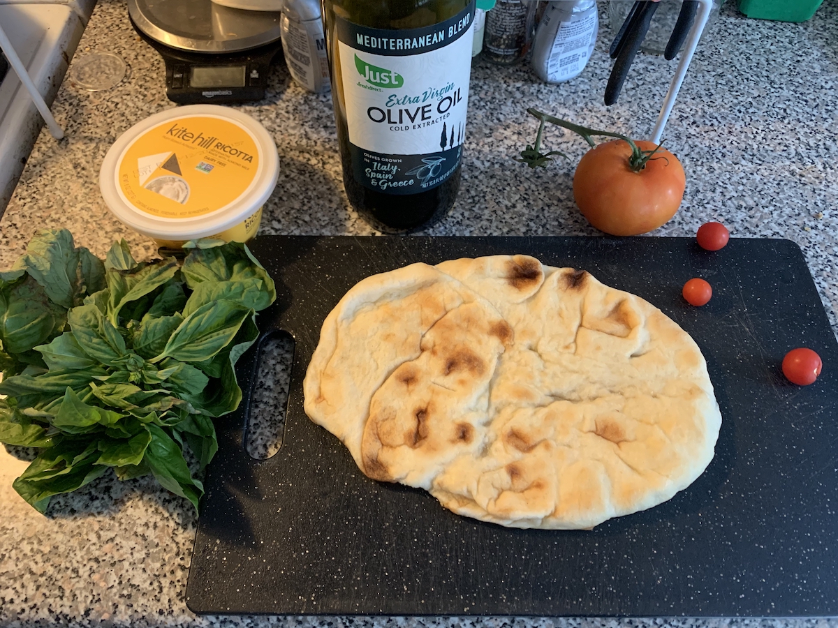 photograph of my counter with ingredients for flatbread: naan, tomatoes, basil, ricotta. they're on a cutting board