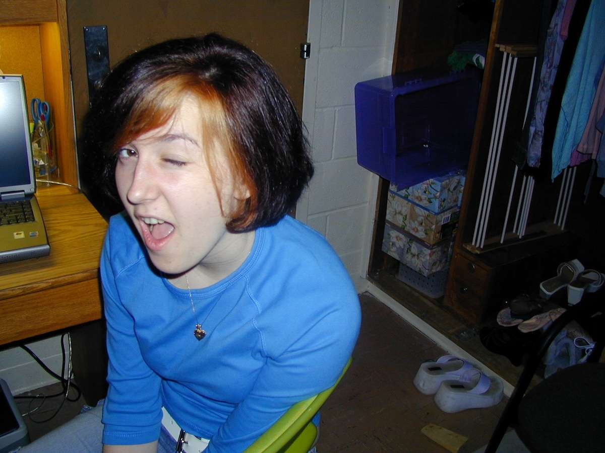photo of me, 2004 at age 19, at my dorm room desk winking. i have a bob with bleached streaks at my side part (professionally done believe it or not, one of my besties was a hair stylist) and i am wearing a white studded belt.